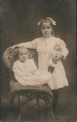 Little Girl Holding a Doll Posing with a Baby Seated on a Chair Postcard