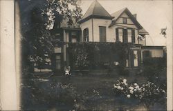 A young boy in a suit is sitting on a bench, holding a baby wearing a gown, behind an old house with a square turret and porch covered in vines. Postcard