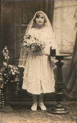 A woman dressed in all white with a white veil poses holding a bouquet of flowers Postcard