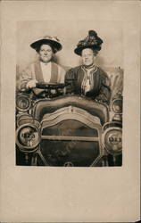 Two women dressed well sitting in the front of a model car Studio Photo Postcard