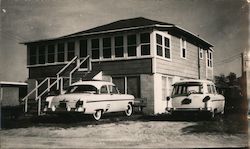 A black and white photo of two antique cars parked outside of a square, two-story house Postcard Postcard Postcard