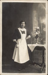 Woman in apron next to flowers on end table Postcard