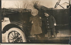 Two small children wearing hats, standing in a parked car. Postcard