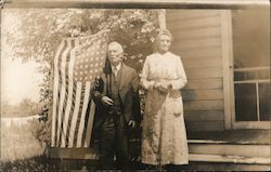 Man and Woman standing by U.S. Flag by porch of house Postcard