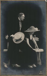 Woman in hat, sitting on a bench; man holding his hat, standing behind her Postcard