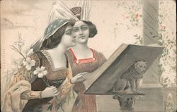 Two women in dresses and hats look at a large book on a stand that bears a carved animal Postcard