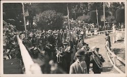 A marching band plays, while a big crowd is gathered for an event. Postcard