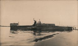 A man raises his hand, while seated on a docked submarine Postcard