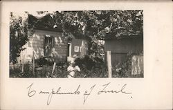 Little girl picking flowers in fenced yard, house, shed Postcard