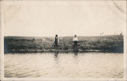 Real photo view of two men standing along water's edge fishing Postcard